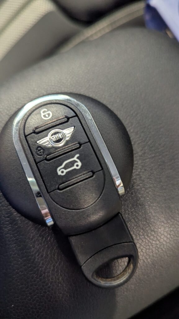 Close-up of a MINI car key fob on a dark surface, showing lock and trunk-release buttons with the MINI emblem in the center.