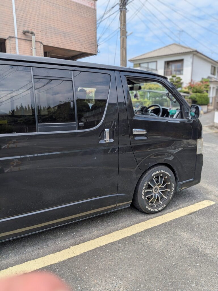 Black van parked along a street with the front door window open; houses and power lines visible in the background.