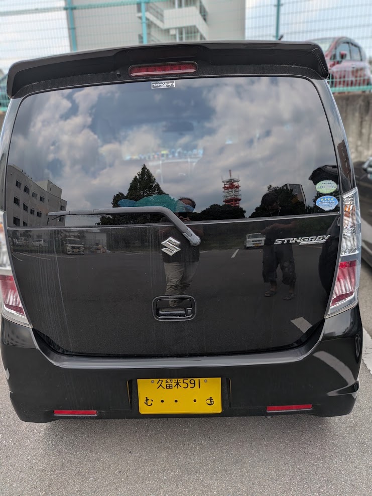Rear view of a black Suzuki Stingray minivan with a yellow Japanese license plate in a parking lot.