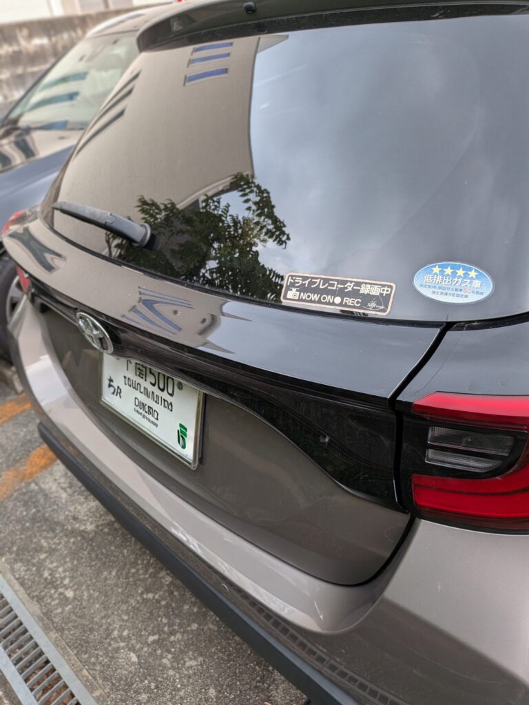 鍵全紛失ヤリス2 - 車専門鍵店カーロック Rear view of a dark SUV with wiper on the rear window and a Japanese license plate at the bottom edge of the tailgate; two stickers on the window (a black'NOW ON REC' label and a blue star badge).