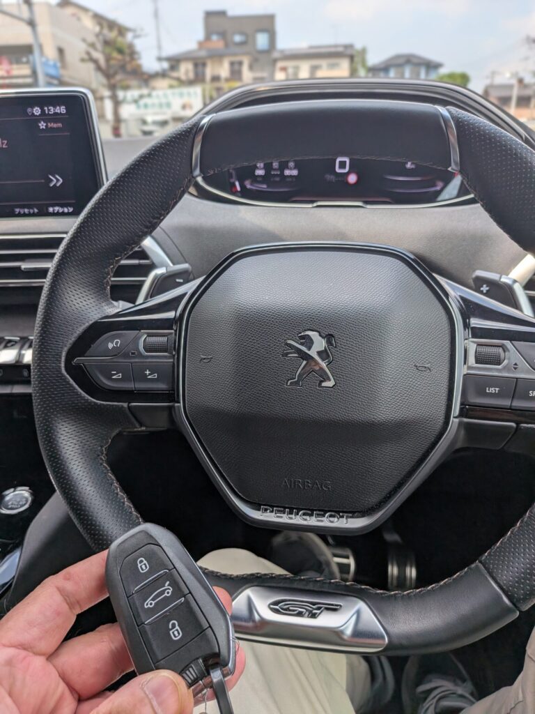 車の車内で、ダッシュボードにGTバッジが見える中、ステアリングホイールの近くでプジョーのキーフォブを手に持っている人物。